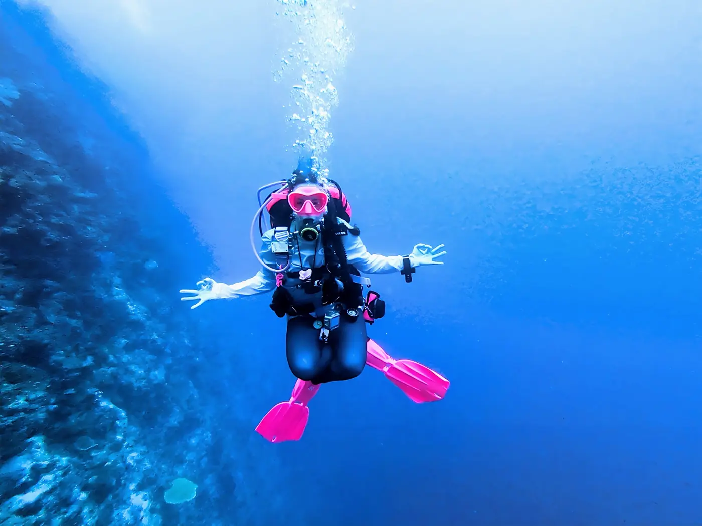 Allie in pink fins giving OK signs beside a reef wall in blue water